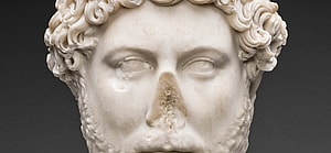 Roman Artifact Portrait Head of Emperor Hadrian, 130-138. The white head of a man with a blank expression, a missing nose that has broken off, low cheekbones, curly hair, and a close-cut wavy beard.