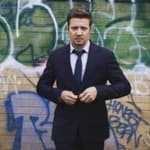 A photo showing Jeremy Renner in a black suit and pant, with a tie to match.