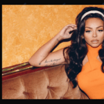 Jesy Nelson sits on a brown couch wearing an orange top and white headband against an orange