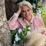 A photo of Dorinda Medley in a pink outfit, leaning against a huge tree in a green field.