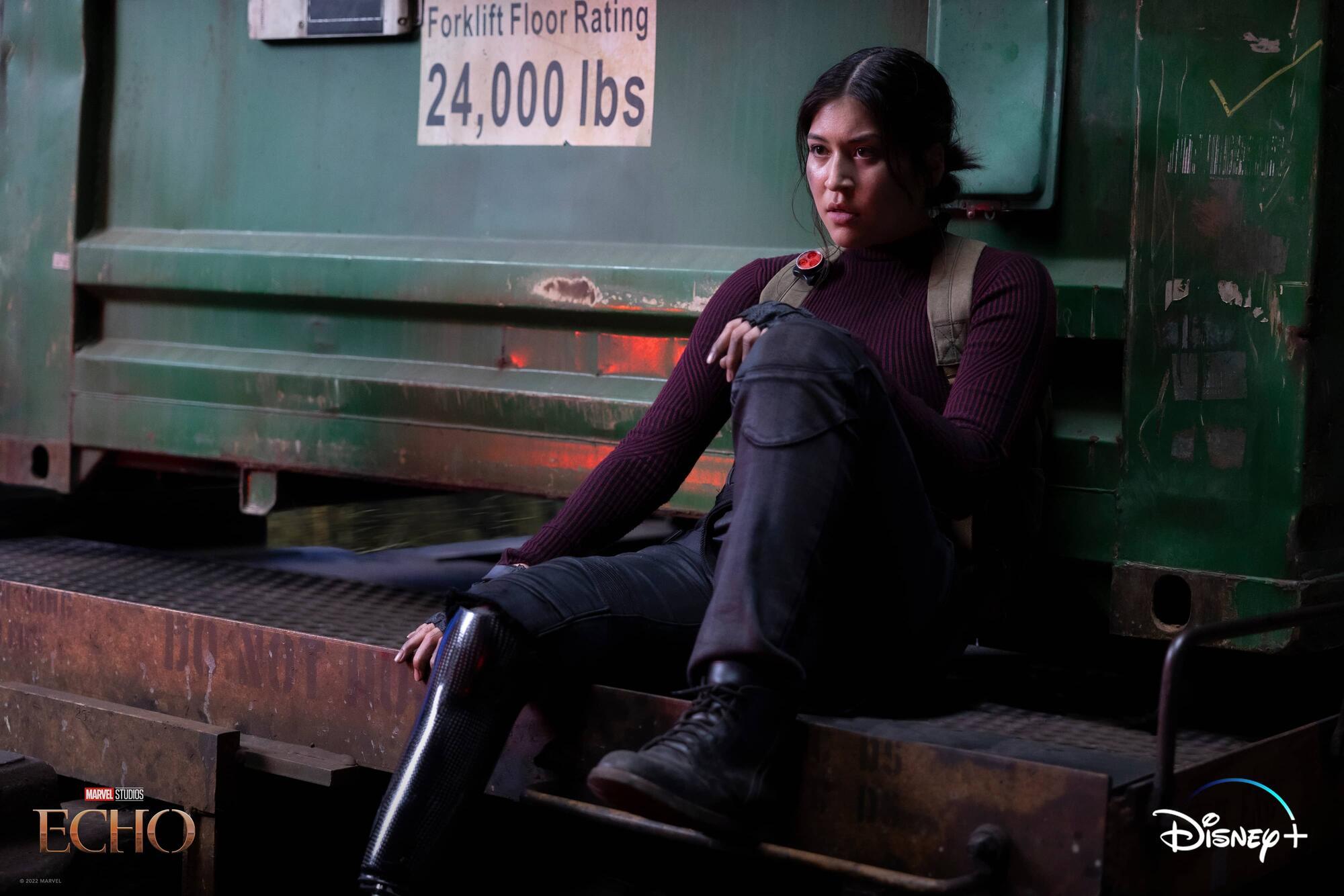 A woman in dark clothing sits on a forklift platform marked "24,000 lbs" in an industrial setting.