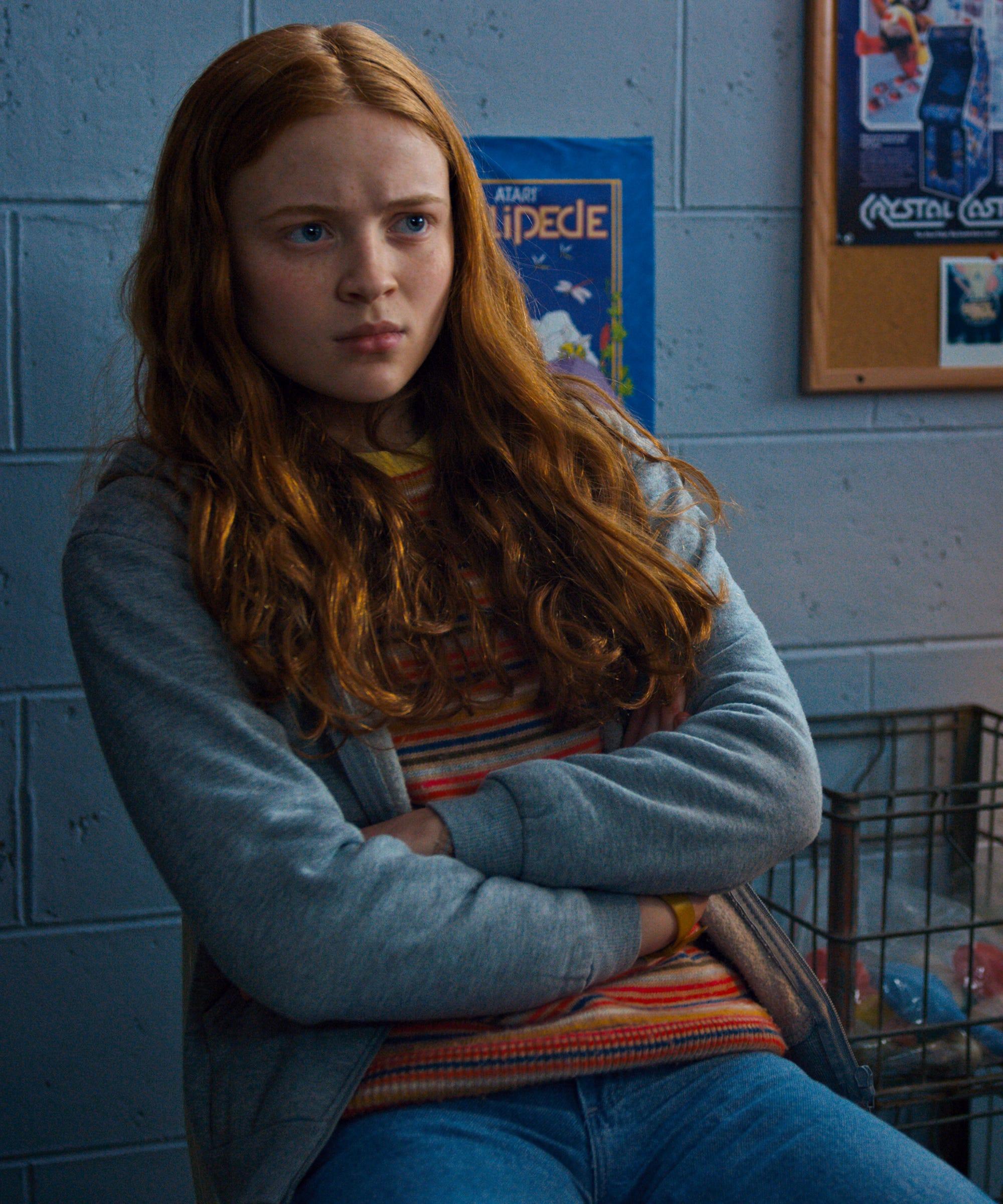 Sadie Sink sitting with arms crossed in a blue-walled room with vintage arcade posters