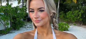 Emily Elizabeth takes a selfie at the beach while wearing a white bikini top and denim shorts.