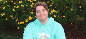 A photo of Shane Dawson in a hoodie, sitting beside a bed of yellow flowers