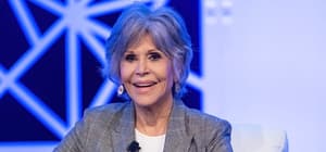 Jane Fonda smiling on stage wearing a plaid blazer at a media event