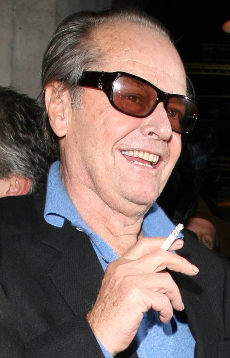 Jack Nicholson wearing sunglasses and a blue shirt, smiling while holding a cigarette