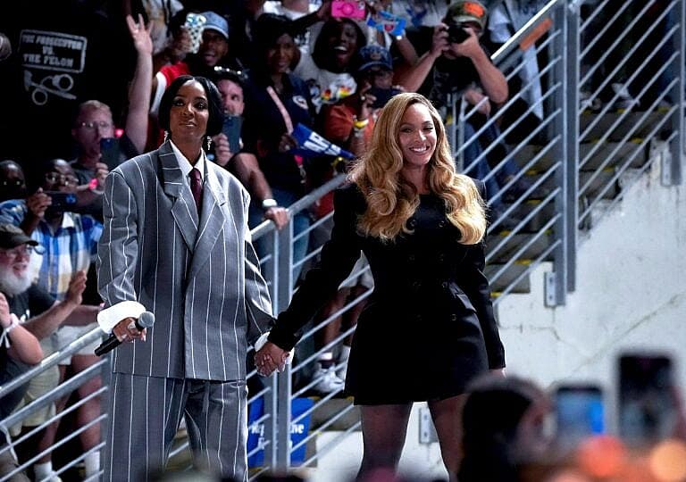 Kelly Rowland and a woman walk down stadium stairs while holding hands, smiling at a crowd.