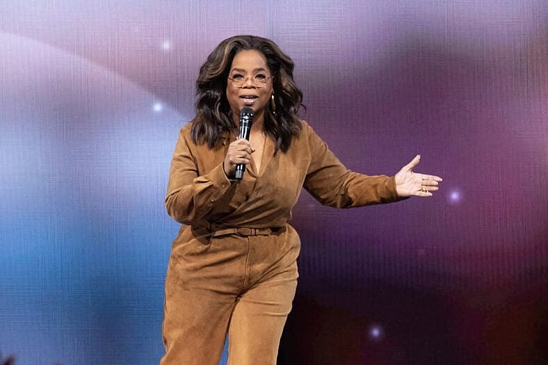 Oprah Winfrey speaking on stage in a tan jumpsuit holding a microphone