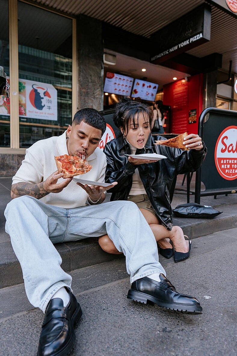 Man and woman sitting on the sidewalk eating pizza.
