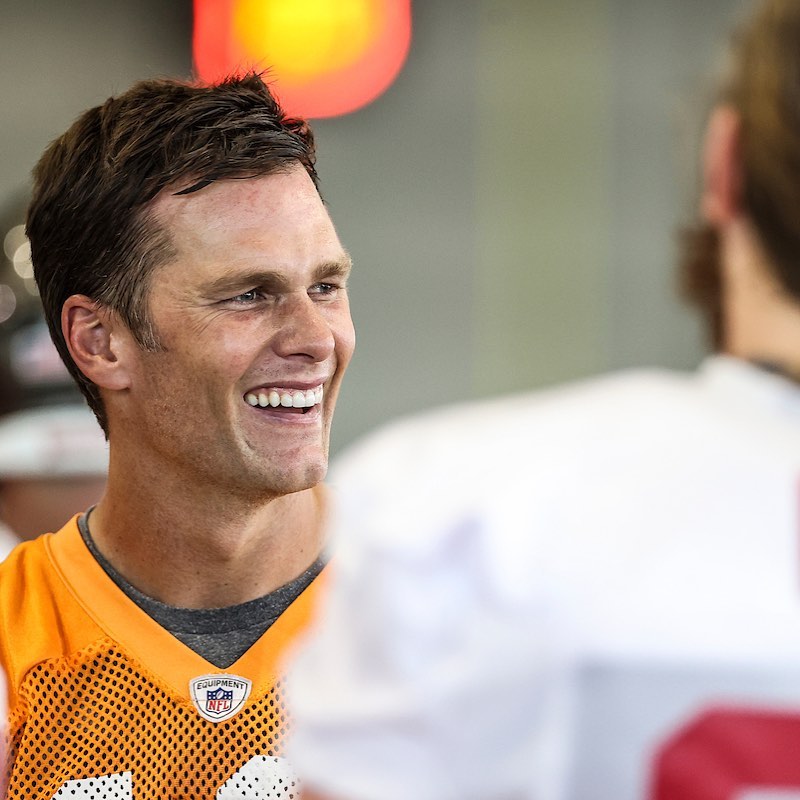 A photo showing Tom Brady in a yellow vest, smiling beautifully.