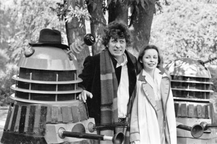Tom Baker poses as the Fourth Doctor in Doctor Who next to a Dalek and Sarah Baker