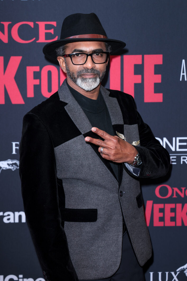 Man posing on red carpet wearing a hat and holding up a peace sign. 