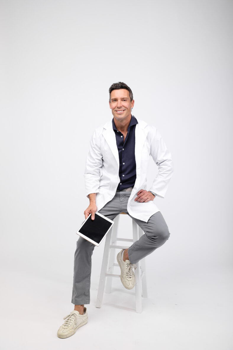 Man in white lab coat sitting on stool.
