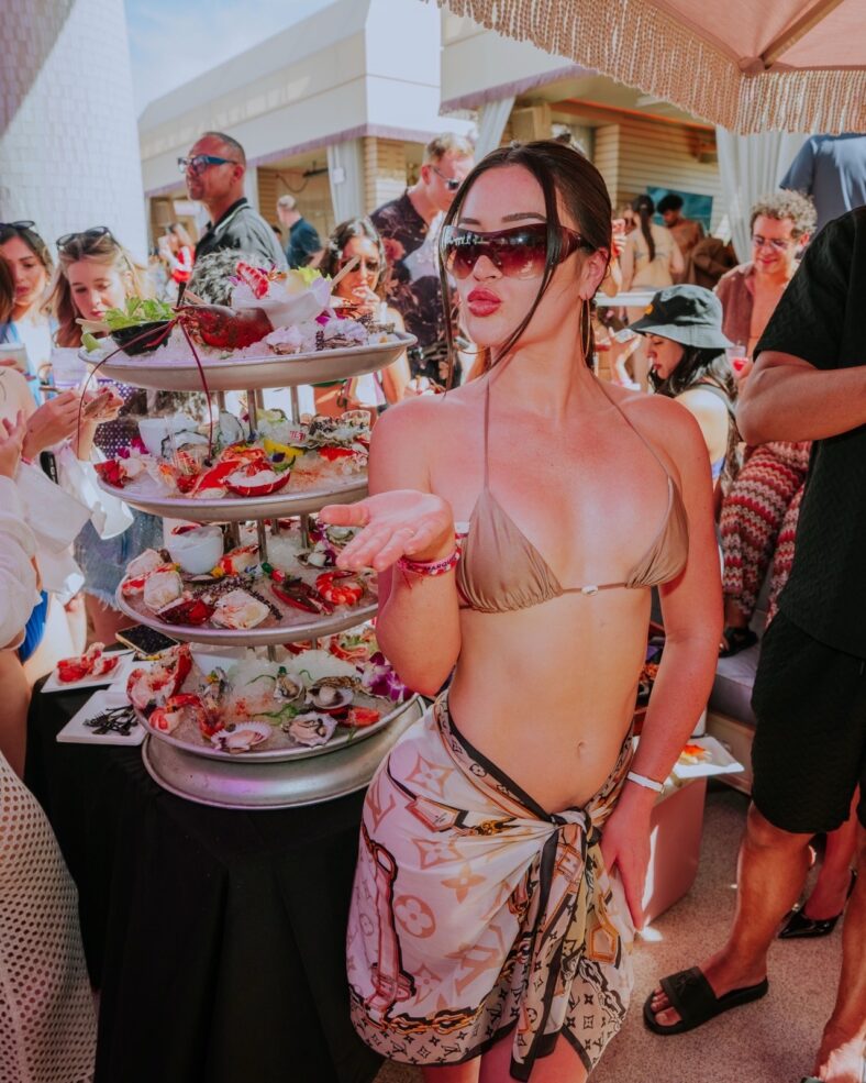 A woman in a bikini blowing a kiss, standing in front of food. 