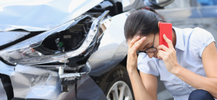 Woman on phone holding head in hands in front of smashed car.