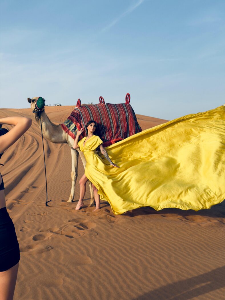 Woman in flowing yellow dress posing in front of a camel in the desert.