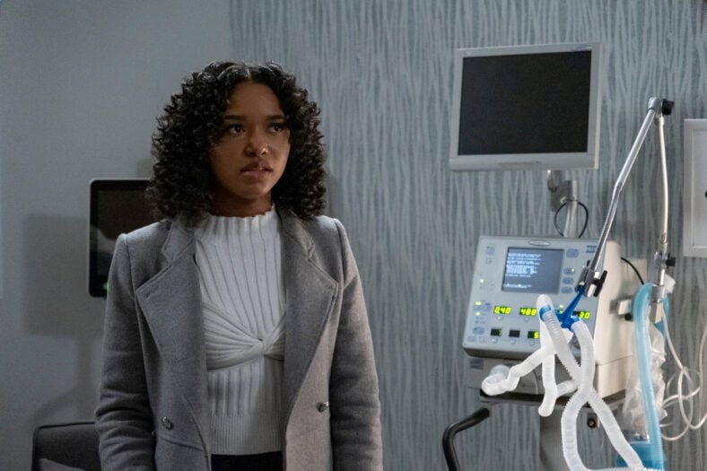 "Bailey Tippen as Sylvie stands in a modern hospital room next to medical equipment and monitors, wearing a gray coat and white sweater, looking concerned."